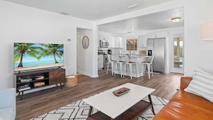 Ein modernes Wohnzimmer mit Fernseher, Couchtisch und Sofa. Im Hintergrund ist eine Küche mit weißen Schränken und ein Essbereich mit weißen Stühlen zu sehen.