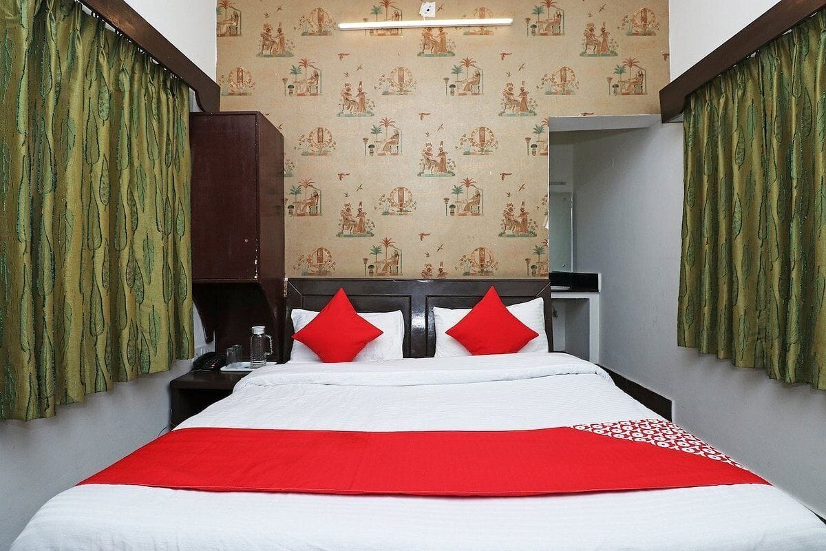 A hotel room with a bed, red pillows, a nightstand, a wooden wardrobe, and patterned wallpaper.