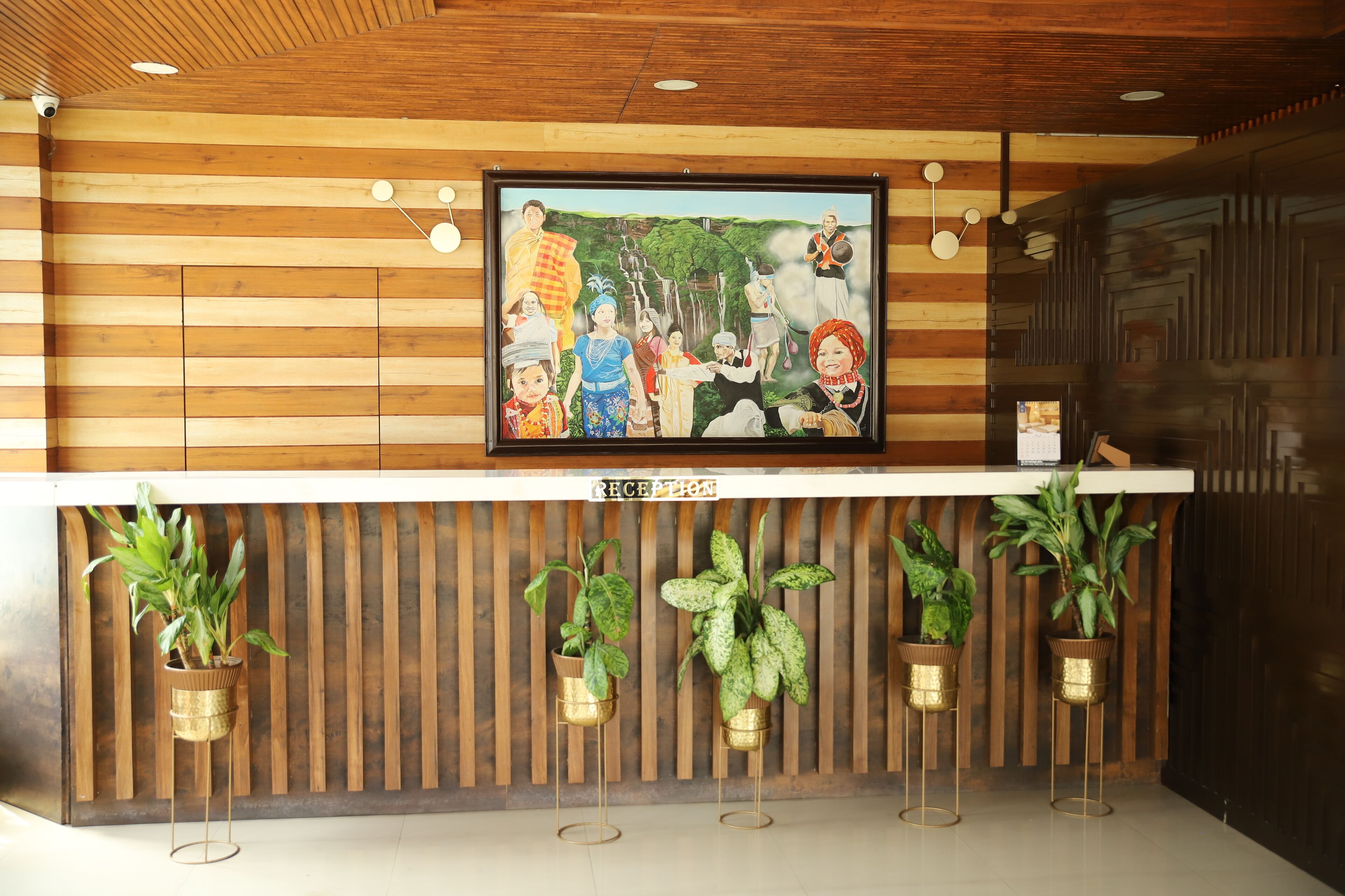 Un comptoir d’accueil avec un panneau décoratif en bois, une œuvre d’art encadrée au-dessus, et des plantes en pot disposées sur des supports.