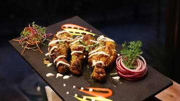 Uma tábua retangular preta com espetos de frango grelhado, uma salada colorida e um enfeite de cebola roxa sobre uma mesa de madeira.