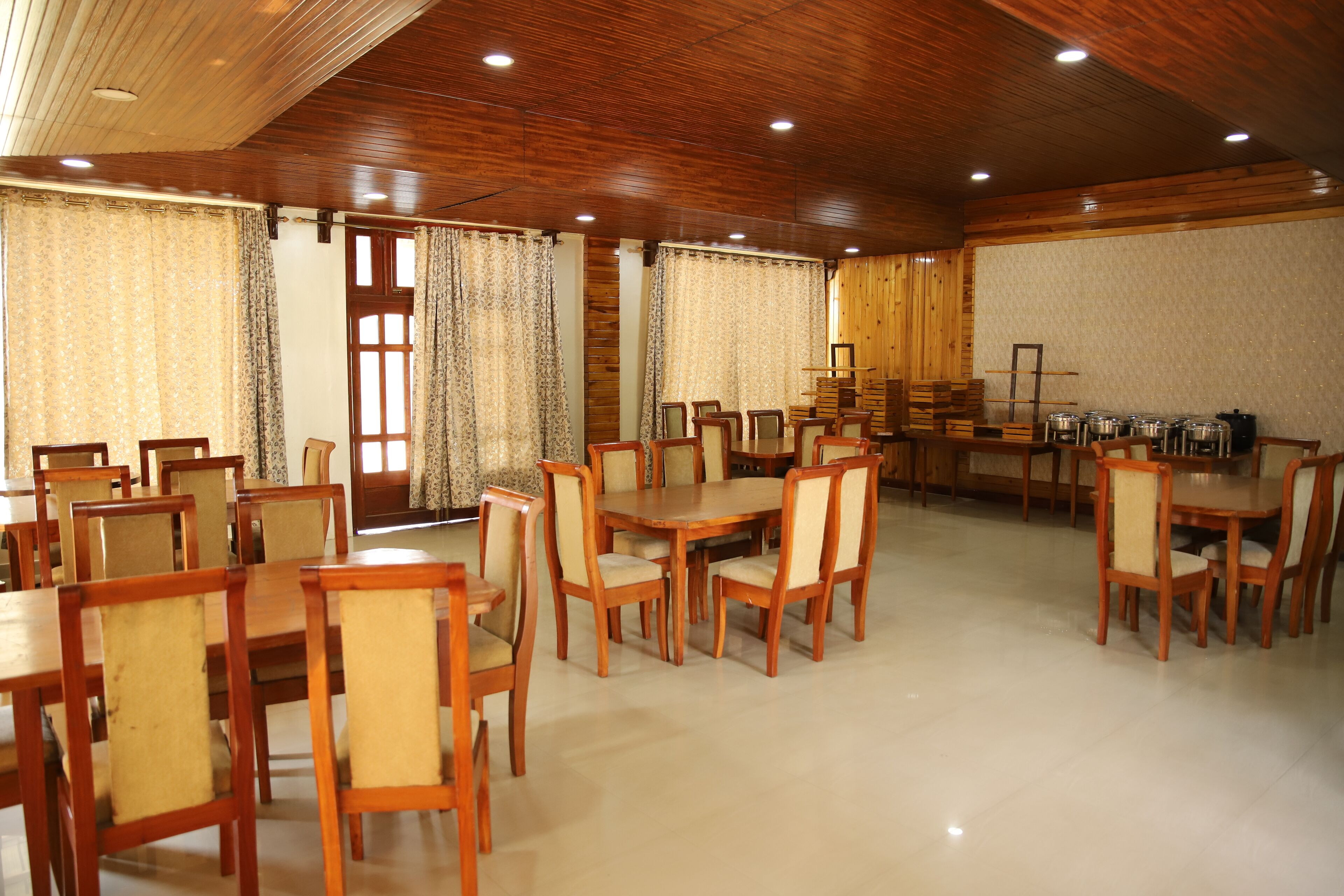 A spacious dining area with wooden tables and chairs, large windows with curtains, and a tiled floor.