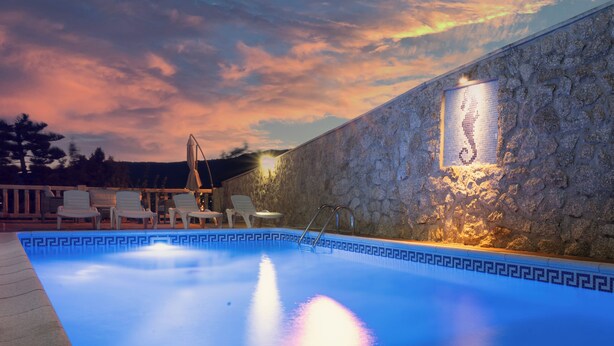 An outdoor swimming pool with a stone wall and a mosaic design, surrounded by lounge chairs and a sunset sky.
