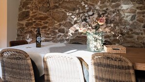 A dining table set with wicker chairs, a bottle of wine, and a vase of flowers against a stone wall.