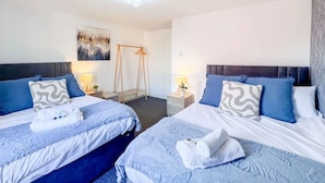 Habitación de hotel con dos camas, cada una con ropa de cama azul y almohadas decorativas. Hay una mesita con una lámpara y una obra de arte colgada en la pared.