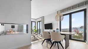 Un salon moderne doté d’une cheminée, d’un coin repas avec deux chaises, d’un grand téléviseur et offrant une vue sur la ville grâce à de grandes fenêtres.