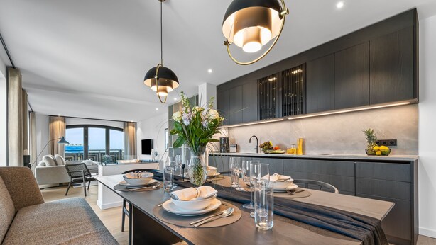 Una cucina moderna con mobili scuri, un'area pranzo con un tavolo lungo apparecchiato per due, e un'ampia finestra con vista sulla città.