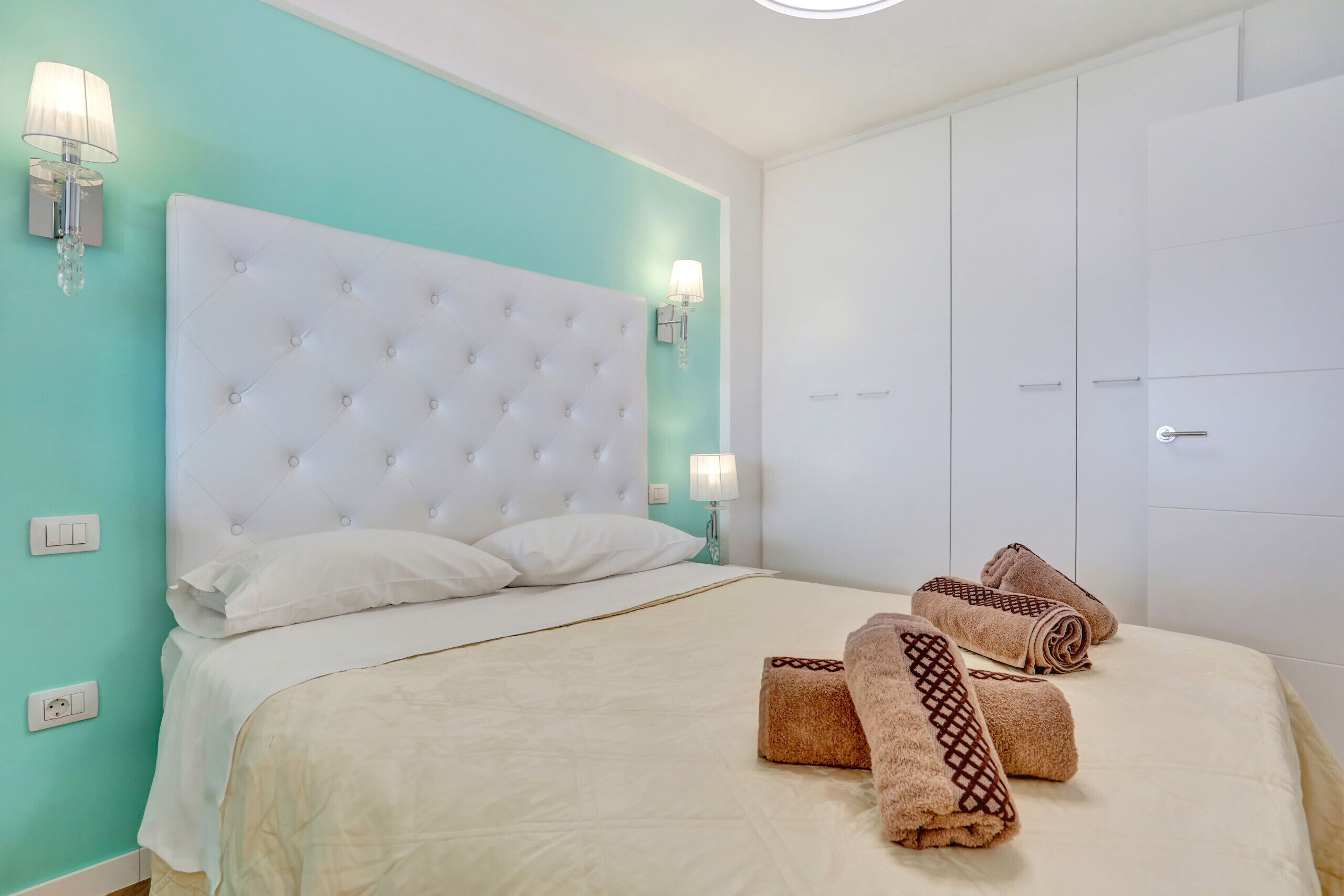 A neatly made bed with a white headboard, two bedside lamps, and rolled towels on the bed.