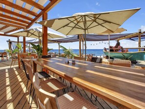 A wooden deck with outdoor seating, including tables and chairs, overlooking a beach with a boat in the background.
