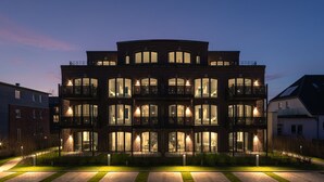 Un immeuble à plusieurs étages, doté de balcons et de grandes fenêtres, entouré d’une pelouse bien entretenue et d’un chemin.