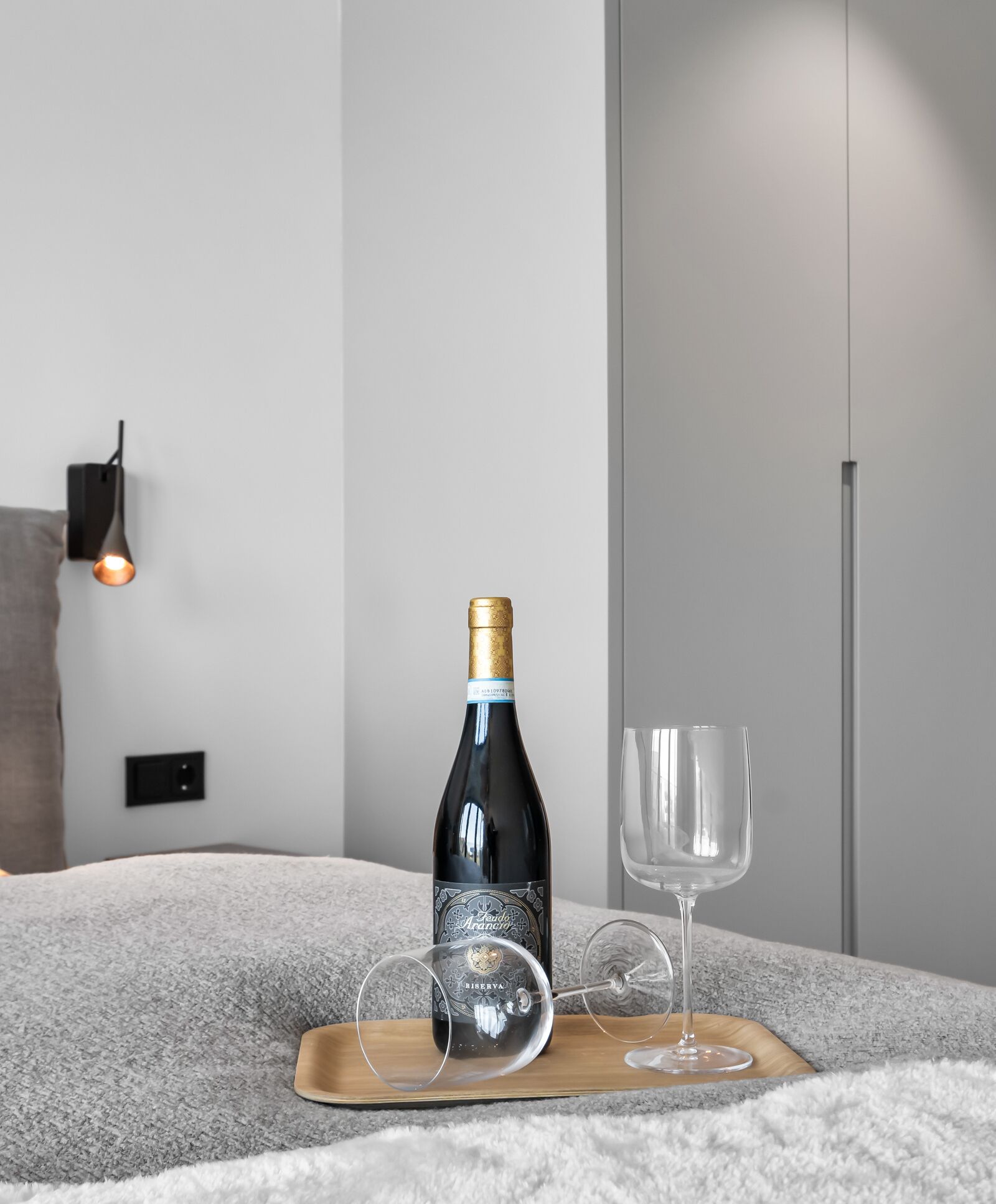 Een fles wijn en een wijnglas op een houten dienblad, gelegen op een grijs tapijt met een structuur.