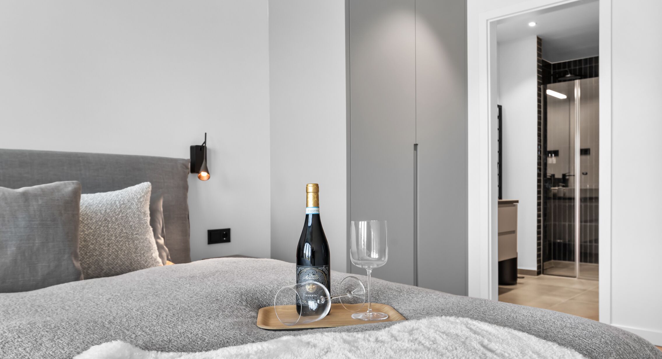 Un lit avec une bouteille de vin et deux verres sur un plateau, une couverture grise et une lampe fixée au mur.
