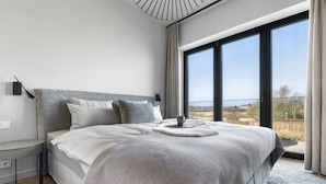 A modern bedroom with a large bed, a bedside table, a lamp, and a view of the outdoors. - Ocean Breeze Suite - Sea View, Wellness, Ethanol Fireplace, Infrared Cabin (List auf Sylt)