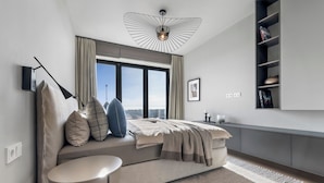 Et moderne soveværelse med et stort vindue, en seng med et beige tæppe, et sengebord, en gulvlampe og en loftlampe.