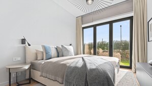 Une chambre moderne avec un grand lit, une table de chevet, une lampe et une vue sur l’extérieur grâce à de grandes fenêtres.
