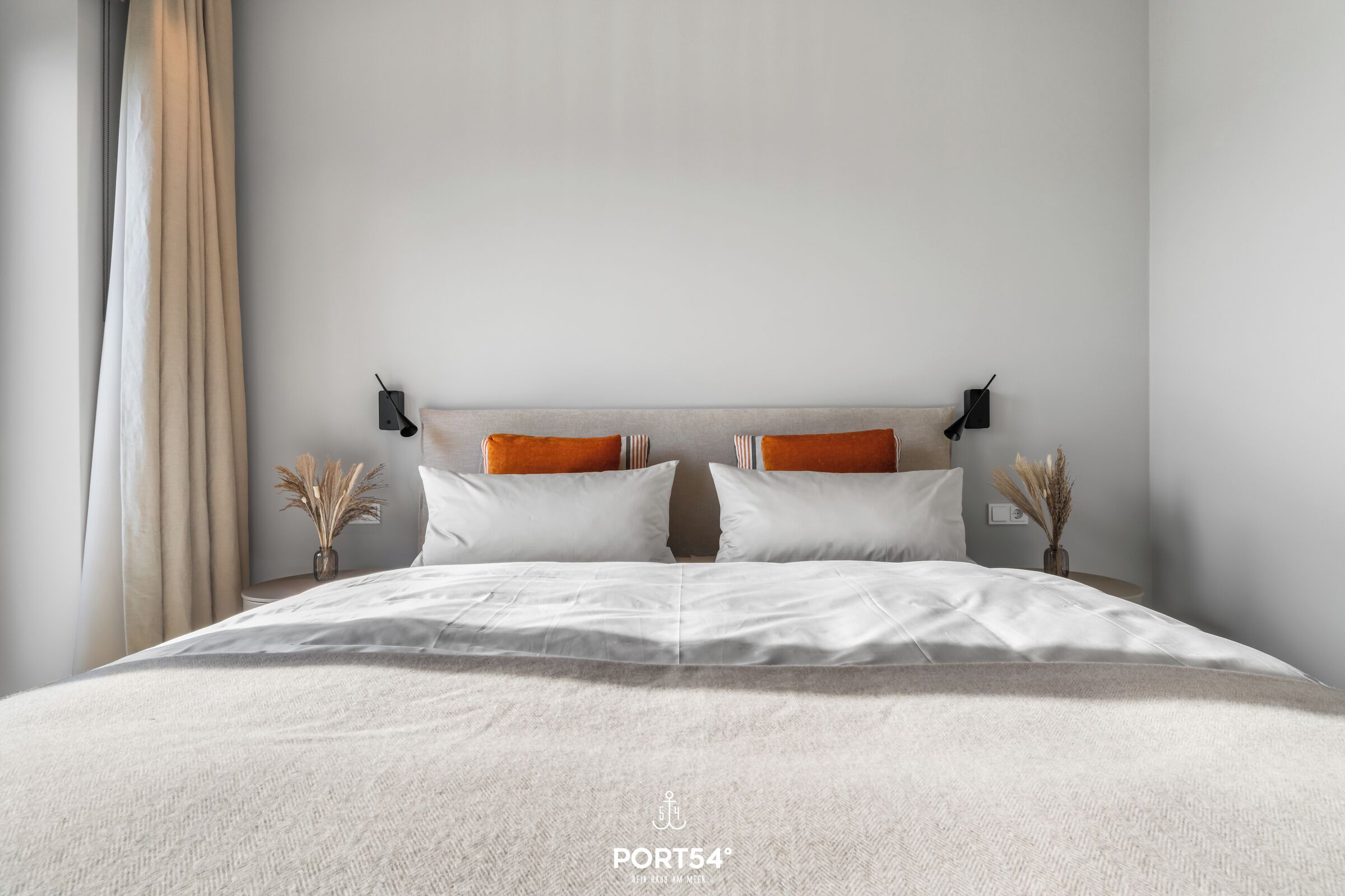 Un lit bien fait, avec du linge de lit blanc et deux coussins orange, une table de chevet ornée d’un vase de fleurs séchées et une lampe fixée au mur.