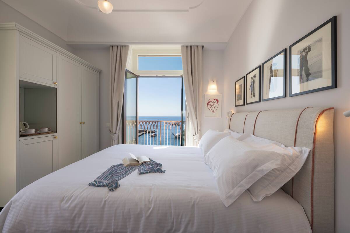 A bedroom with a large bed, a view of the sea through a balcony, and a set of framed pictures on the wall.