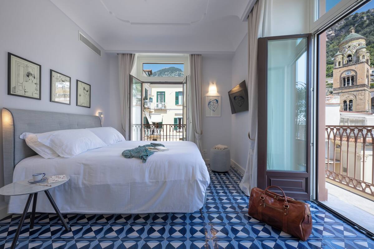 A hotel room with a bed, a table with a cup and a remote, a window with a view of a building, and a patterned floor.
