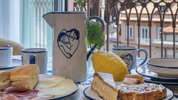 A table with food items and a pitcher with a heart design, overlooking a historic building and greenery. - ABBRACCIO Amalfi Boutique Retreat (Amalfi)