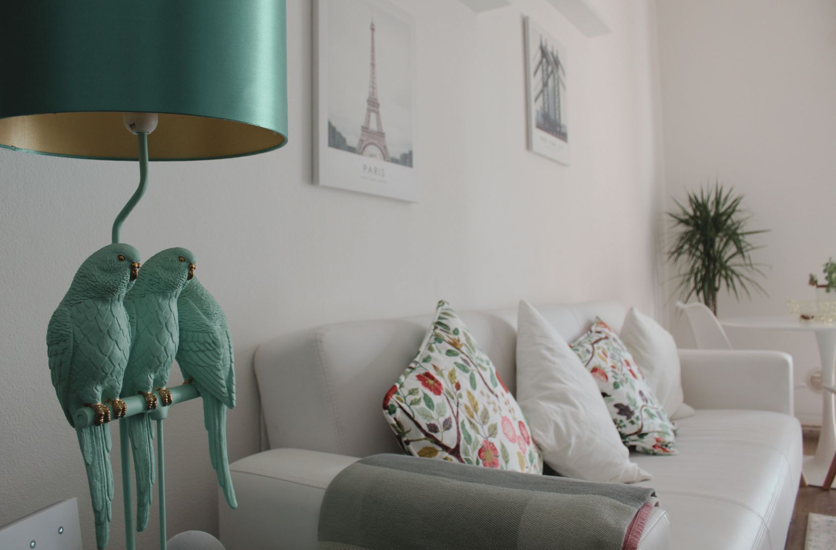 Una sala de estar con un sofá blanco, guacamayos verdes en un soporte, una lámpara verde azulada y cuadros en la pared.