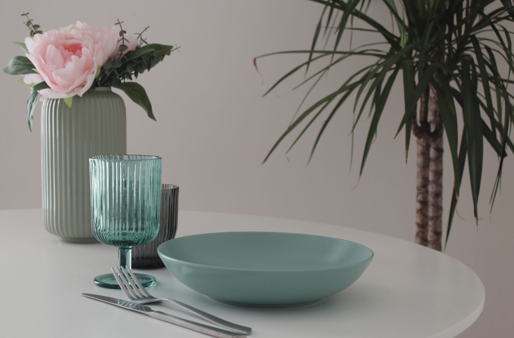 Una mesa de comedor con un tazón turquesa, un vaso y cubiertos. Un jarrón con una flor rosa y hojas verdes está sobre la mesa. Una planta alta con hojas largas se ve en el fondo.