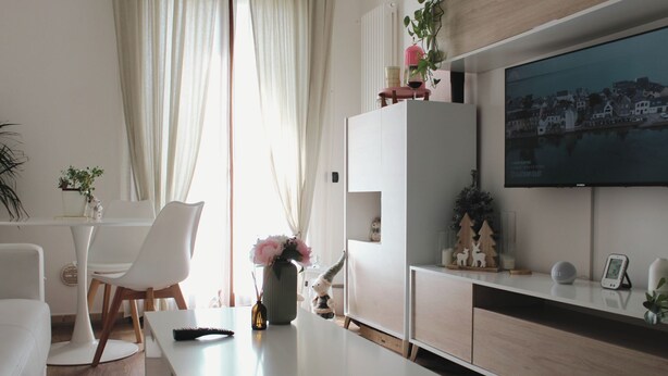 Un salon moderne avec un canapé blanc, une table basse et un téléviseur fixé au mur. On trouve une plante sur la table basse et une petite étagère ornée d’objets décoratifs.