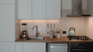 Une cuisine moderne avec des meubles blancs, un évier en acier inoxydable, un mixeur, une bouilloire et une cuisinière à gaz.