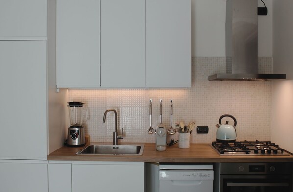 A modern kitchen with white cabinets, a stainless steel sink, a blender, a kettle, and a gas stove.