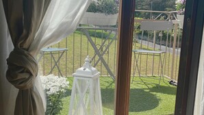 Une vue depuis une fenêtre dont le rideau est retenu, dévoilant un balcon avec une lanterne blanche, une petite table et une balançoire.