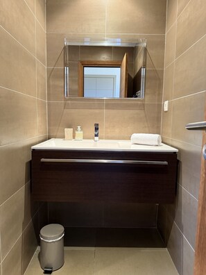 Une salle de bain moderne avec un meuble-lavabo en bois foncé, un lavabo blanc et un miroir au-dessus. On y trouve une poubelle et une serviette sur le comptoir.