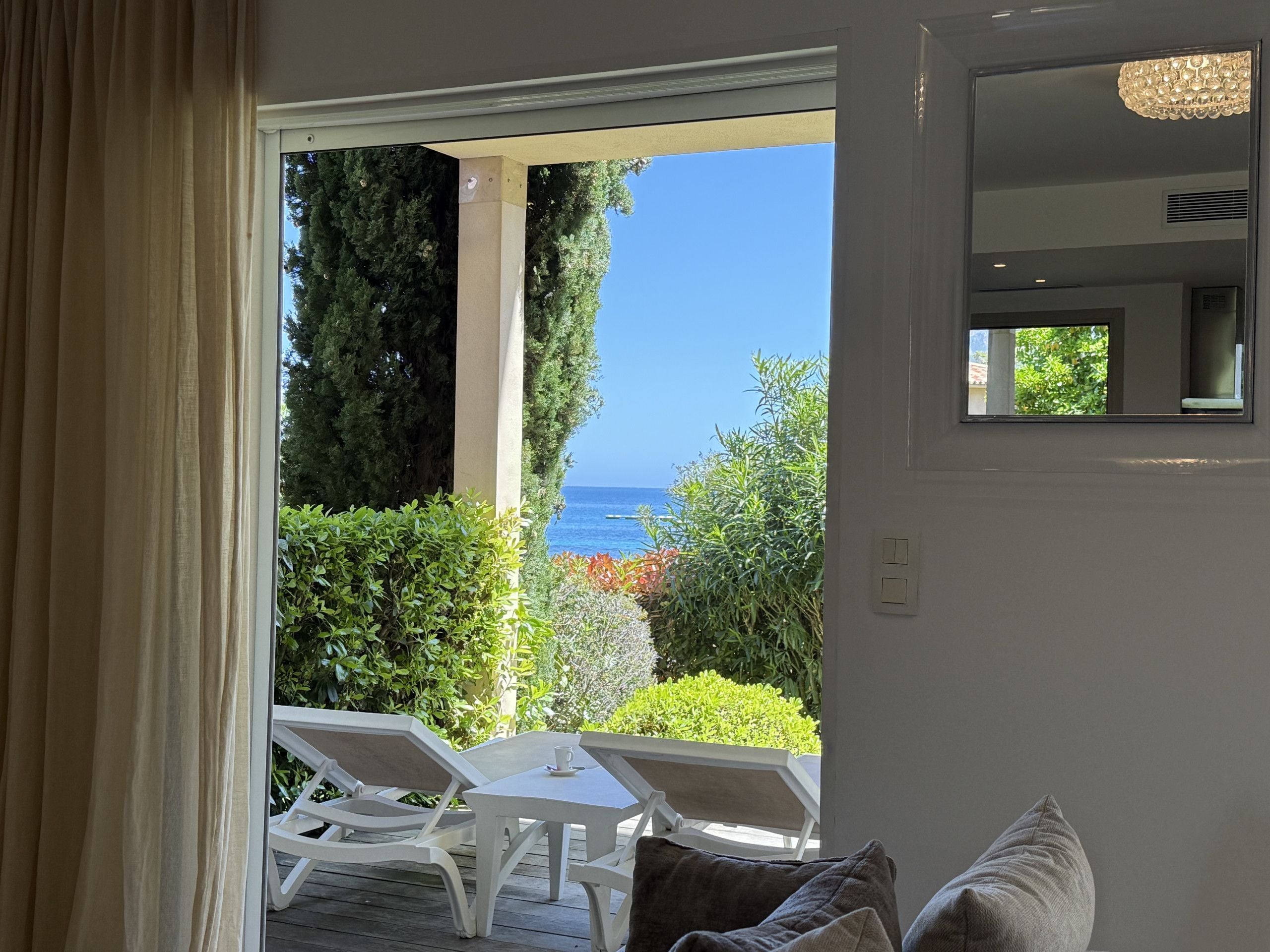 A room with a view of the sea, a white table with chairs, and a window with a view of greenery.