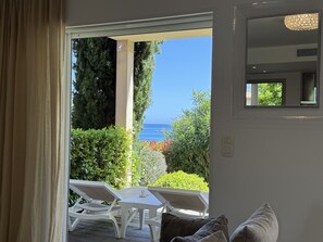 A room with a view of the sea, a white table with chairs, and a window with a view of greenery.