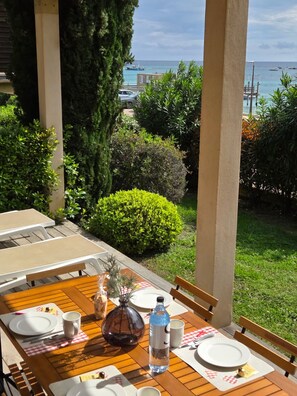 Une table en bois, dressée pour deux personnes, avec des assiettes, des tasses et une bouteille, donnant sur un jardin et la mer.