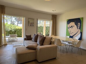 A modern living room with a sectional sofa, a dining table with chairs, large windows, and a painting on the wall.
