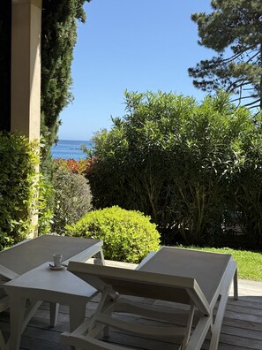 Een terras met twee witte tafels en stoelen, een klein wit kopje op een tafel, en zicht op zee en groen op de achtergrond.