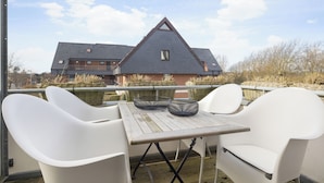 Un balcon avec une table et deux chaises blanches, donnant sur une maison dont le toit est triangulaire.
