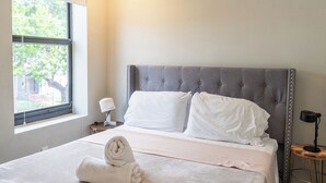 A neatly made bed with a grey upholstered headboard, a bedside lamp, and a small wooden table with a lamp. - Trendy Brickell 2bd/2bth Free Parking Apt 5 (Miami)