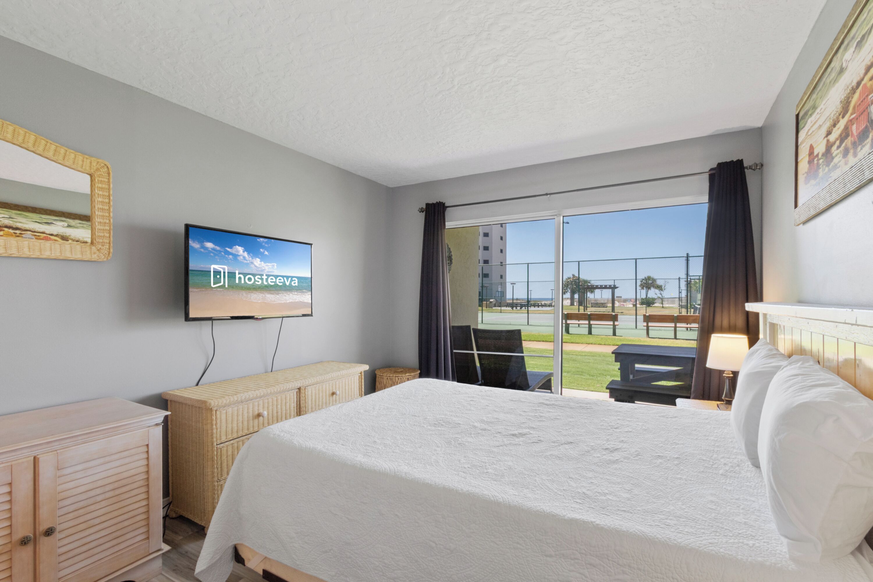 Habitación de hotel con cama, televisor, cómoda de mimbre y vistas al exterior.