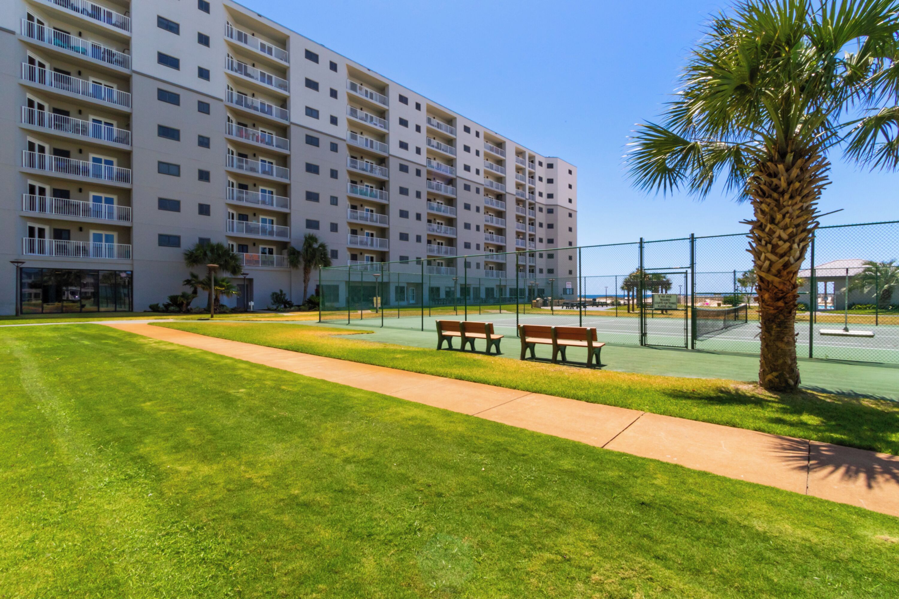 A multi-story hotel with balconies, a tennis court, and a palm tree.