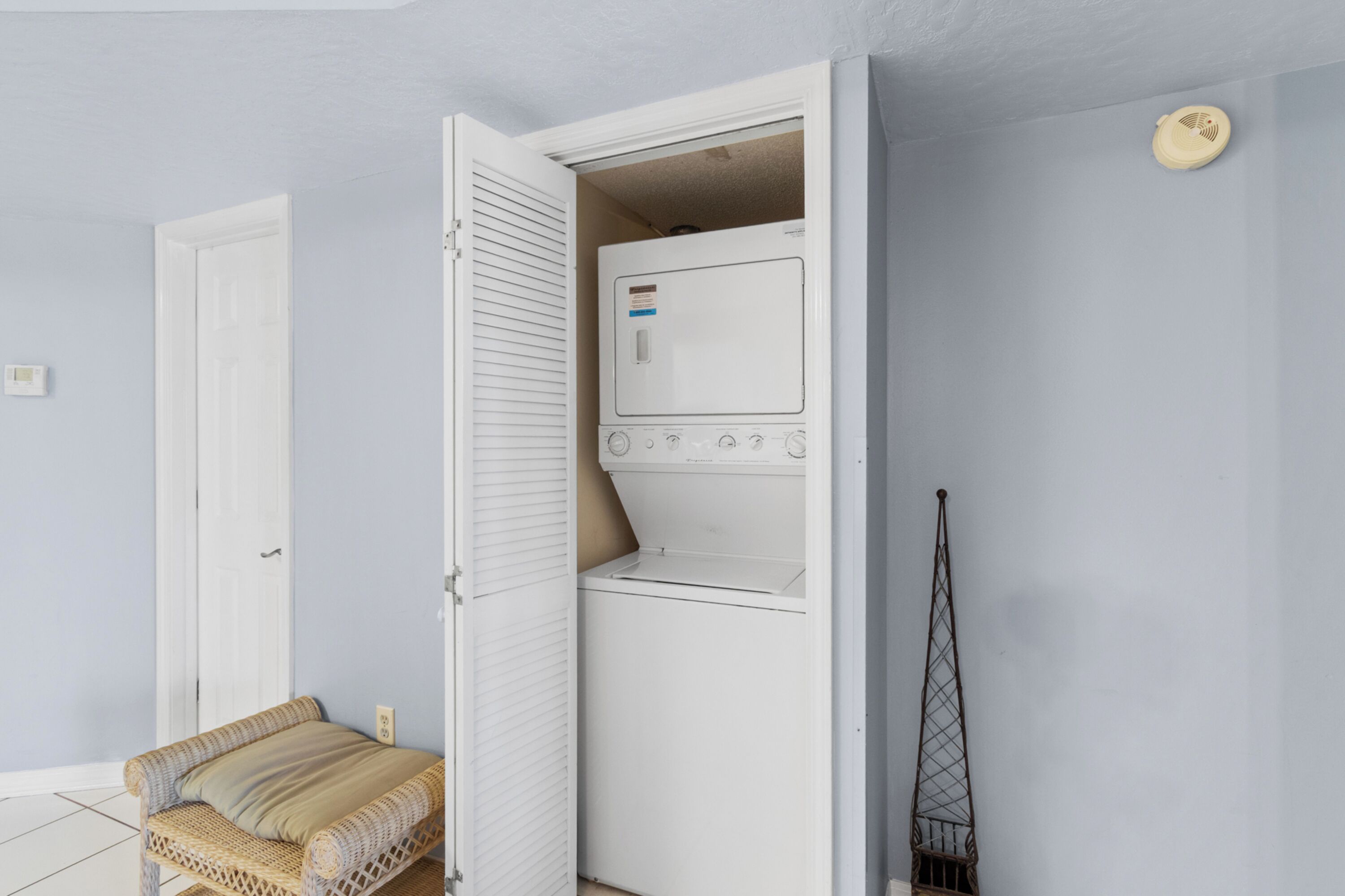 Uma lavanderia com máquina de lavar e secadora, uma cadeira de vime e uma porta.