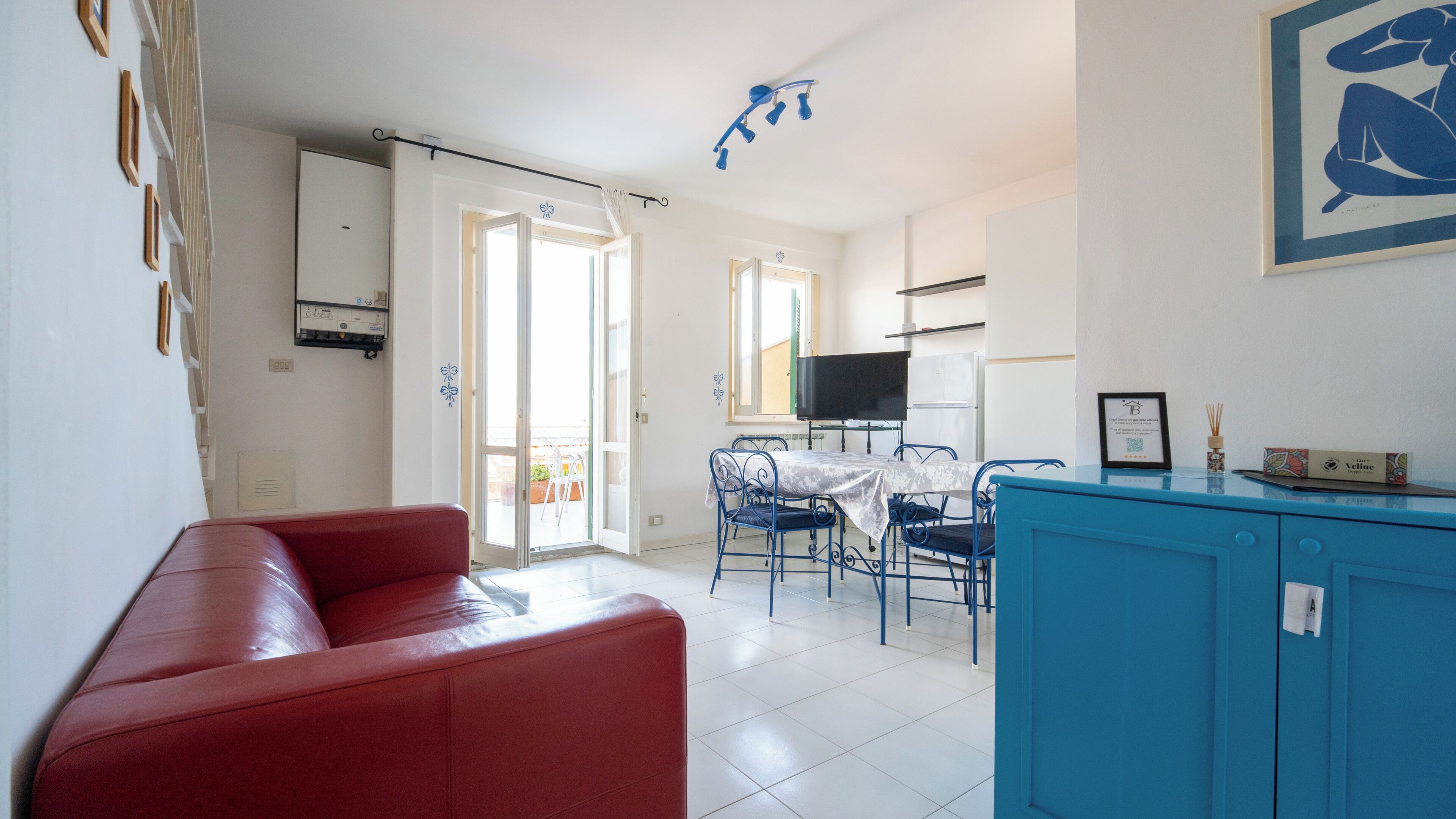 Un salon avec un canapé rouge, un coin repas avec une table et des chaises, une cuisine avec un téléviseur et un balcon avec des chaises.