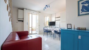 Un salon avec un canapé rouge, un coin repas avec une table et des chaises, une cuisine avec un téléviseur et un balcon avec des chaises.