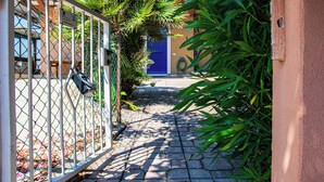 Un étroit sentier pavé mène à un bâtiment doté d’une porte bleue, entouré d’une végétation luxuriante et d’un portail en métal.