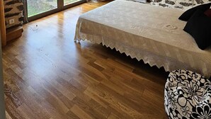Une chambre avec un mur en pierre, des poutres en bois, un lit recouvert d’une housse en dentelle, un radiateur et une fenêtre donnant sur un paysage verdoyant.
