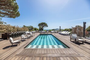 Une piscine rectangulaire à l’eau d’un bleu cristallin, entourée d’une terrasse en bois et de chaises longues blanches.
