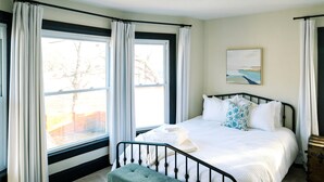Un dormitorio con cama, un banco, una ventana con cortinas y un cuadro en la pared.