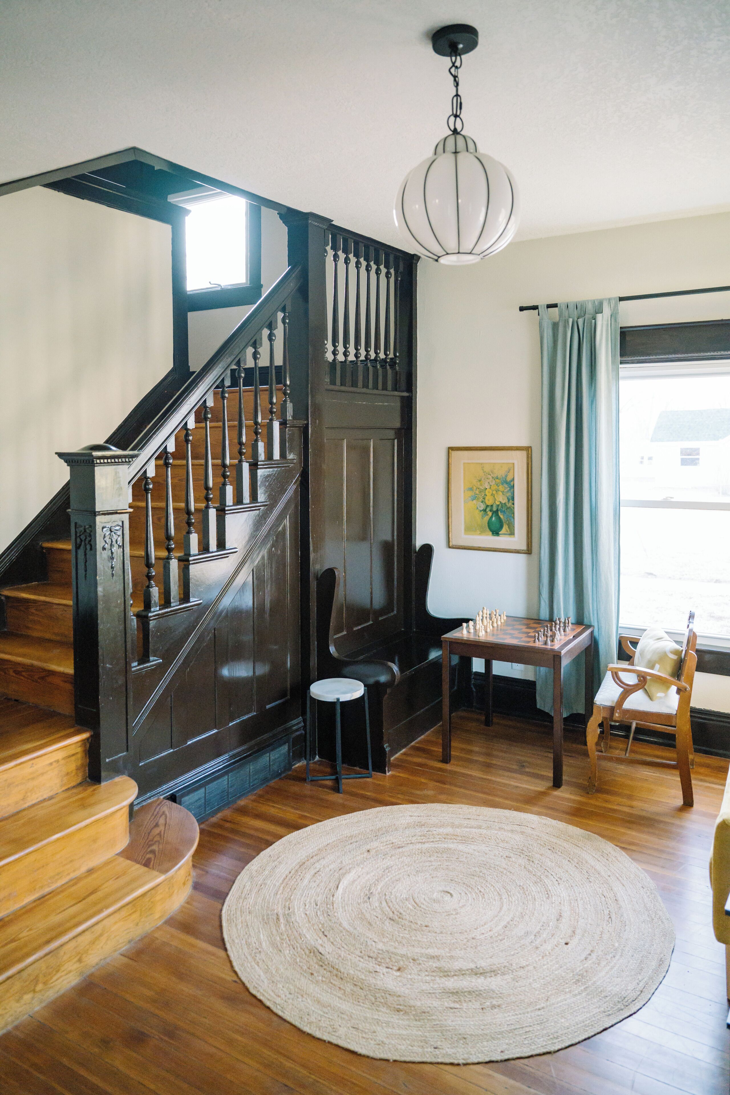 Una escalera de madera con un candelabro, un tablero de ajedrez y una alfombra redonda en una habitación con una ventana y un cuadro.