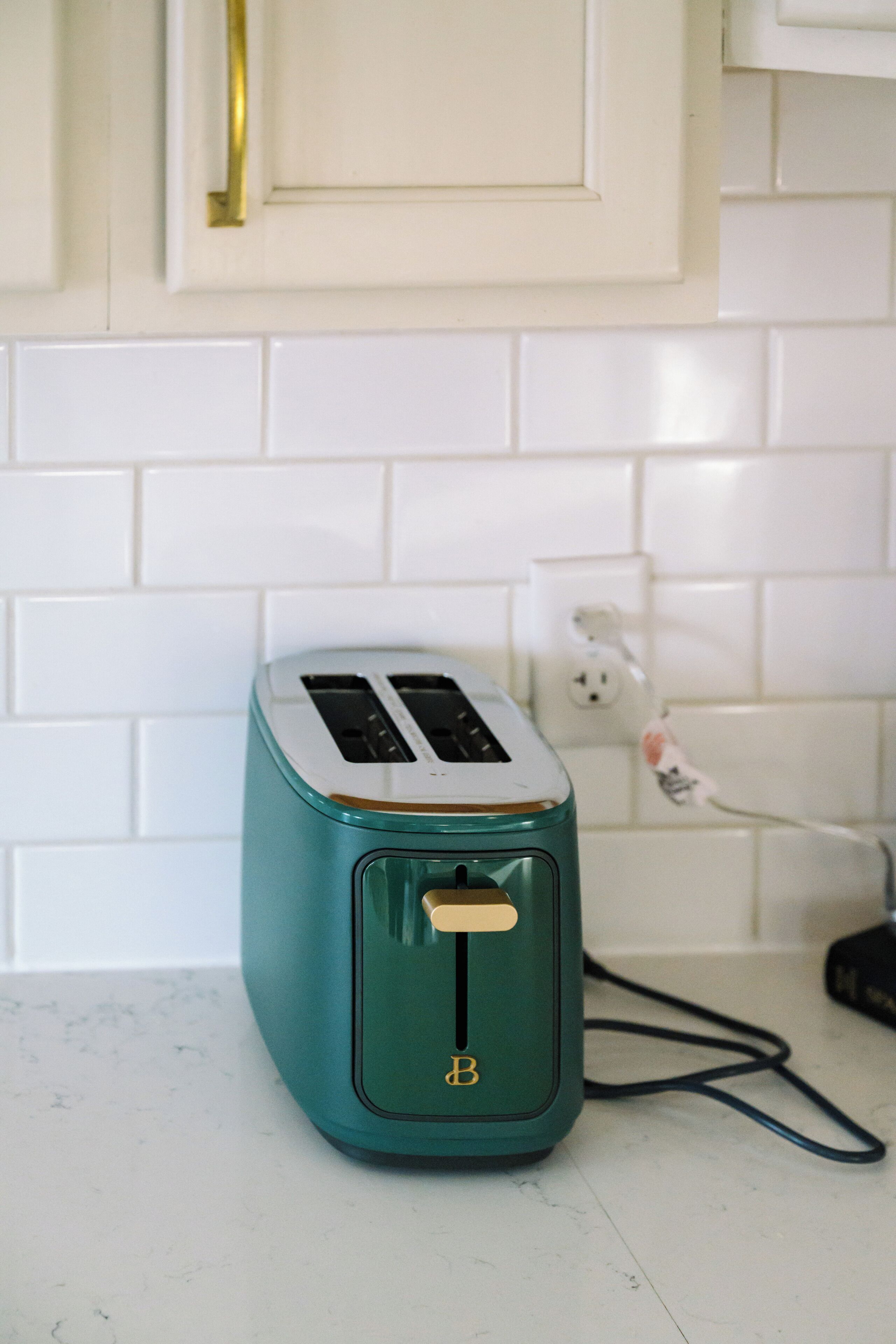 Een blauwgroene toaster op een wit aanrecht met een witte betegelde achterwand en een goudkleurige hendel erboven.