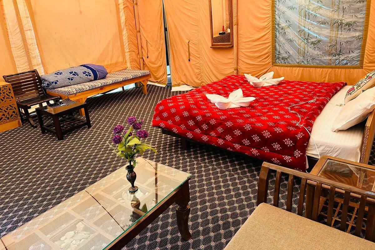 A room with a bed, a sofa, a wooden chair, a glass coffee table, and a small table with a vase.
