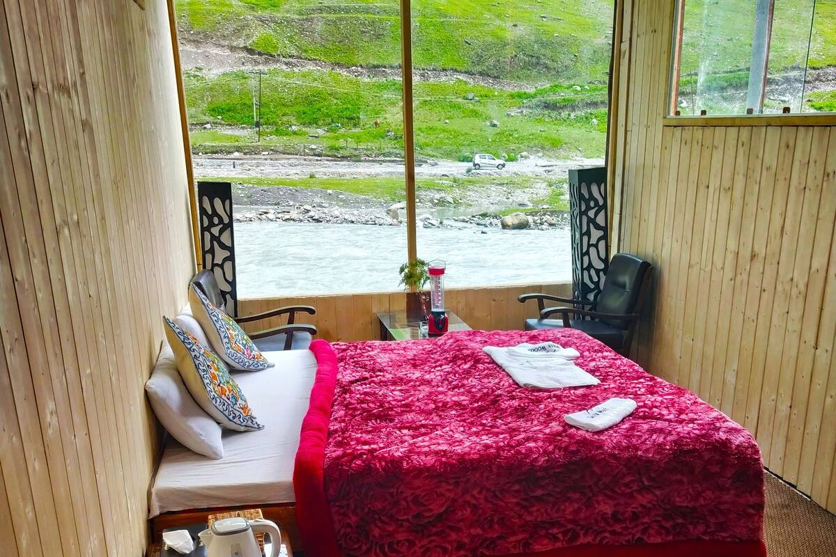 A room with a bed, a rocking chair, a table with a kettle, and a view of a river and green landscape.