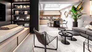 Un salon moderne avec un canapé, une table basse et un fauteuil. On aperçoit une cuisine au fond, avec une table à manger et des chaises.